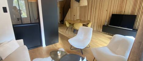 A modern living room with a sofa, two armchairs, a coffee table, and a dining area with a table and chairs. A flat-screen TV is mounted on the wall.