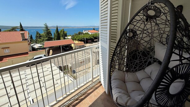 A balcony with a hanging chair, a view of the sea, and buildings in the background. - Apartment Val (Crikvenica)