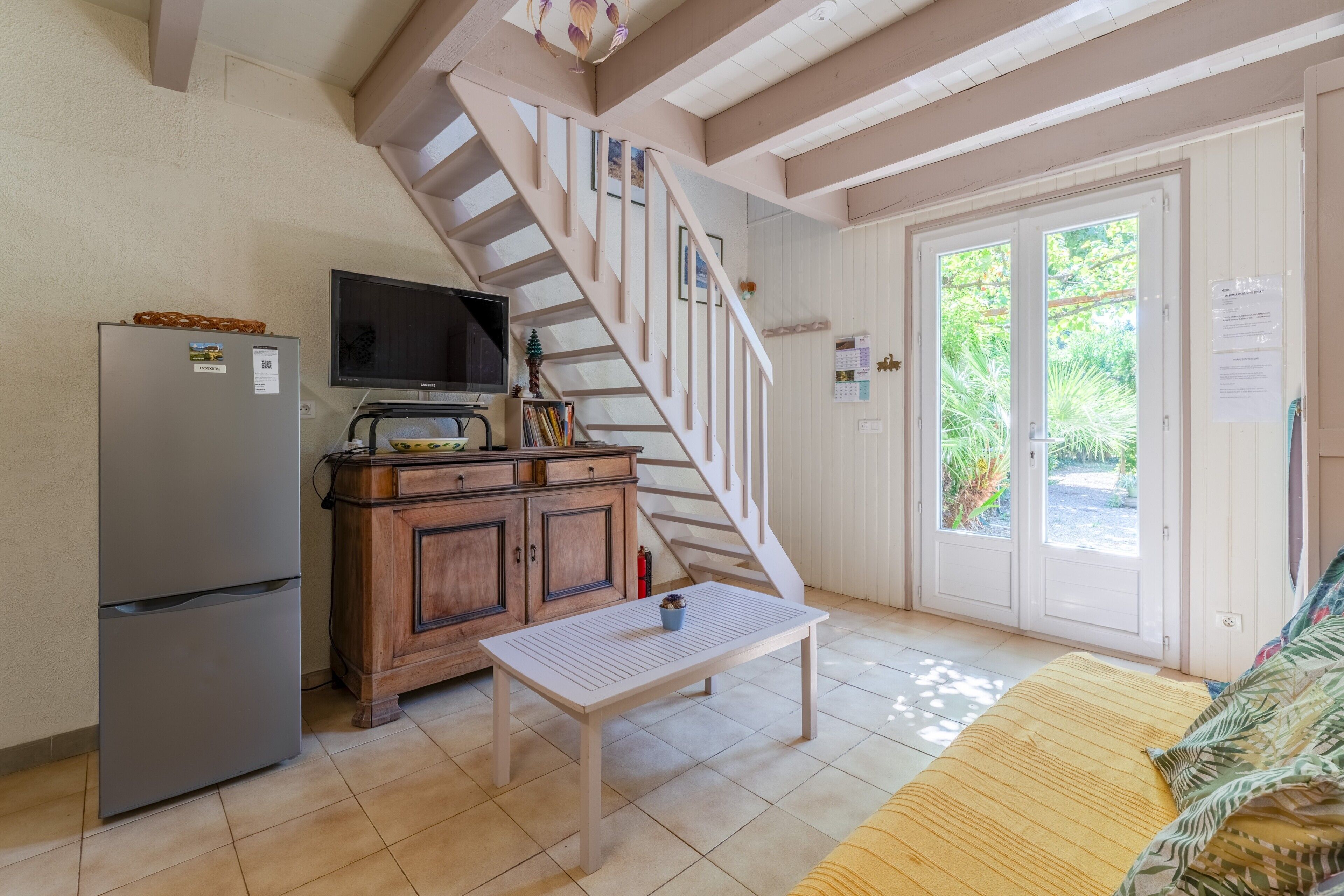 A kitchen with a refrigerator, wooden cabinets, a television, a table with a bowl, and a staircase leading to a door with a view of greenery outside.