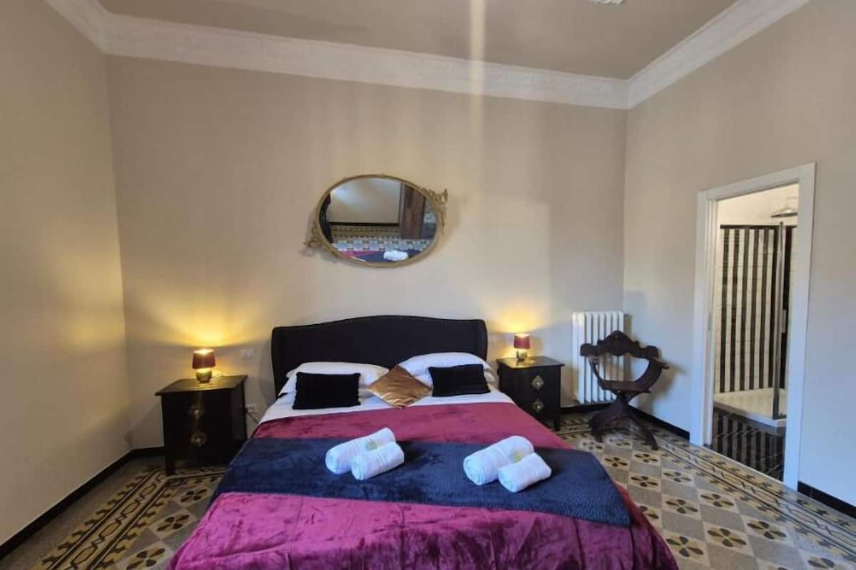 A bedroom with a bed, two bedside tables with lamps, a round mirror, and a bench.