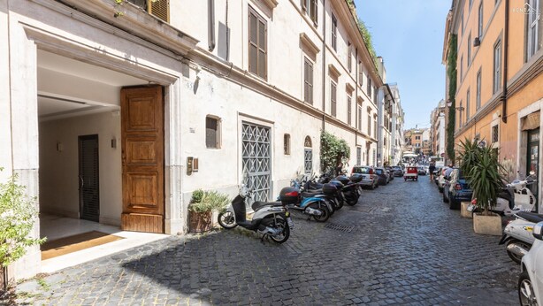 A cobblestone street lined with parked scooters and cars, leading to a building with a wooden door and a metal gate.