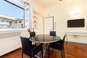 Un comedor moderno con una mesa redonda de vidrio, sillas negras, un armario blanco con estantes, una puerta y un televisor montado en la pared.