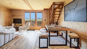 Ein gemütliches Blockhaushaus-Interieur mit Holzbalkendecke, einem großen Fernseher, einem Essbereich und einer Treppe.