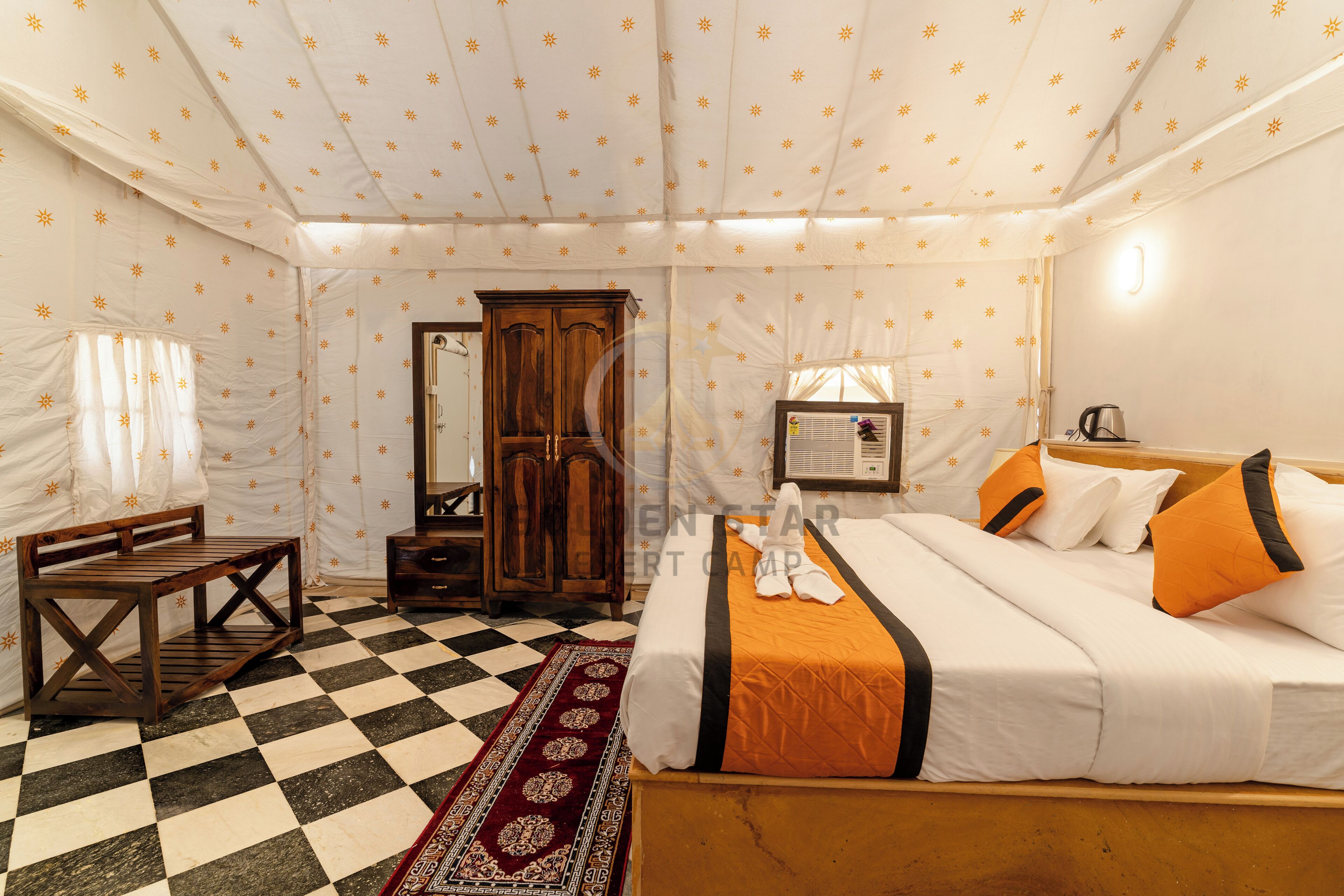 Een hotelkamer met een geruite vloer, een bed met wit en oranje beddengoed, een houten bankje, een kledingkast en een kamerbreed tapijt met een patroon.