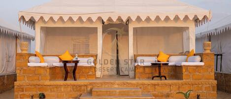 A desert camp with a tented structure, two sofas with cushions, a table, and potted plants.