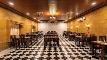 Um quarto com chão xadrez, cadeiras e mesas de madeira, um candelabro e luminárias fixadas na parede.