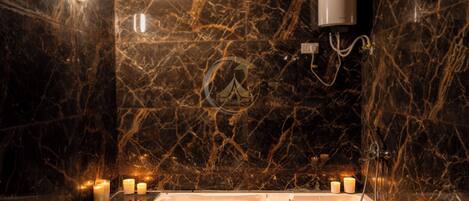 A bathroom with a marble wall, a bathtub filled with rose petals, and several lit candles.