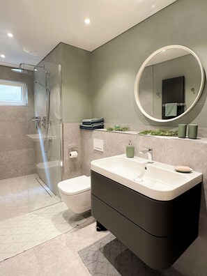 Un baño moderno con ducha envuelta en vidrio, inodoro blanco, tocador grande con espejo y lavamanos pequeño.