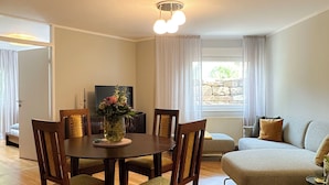 Ein Essbereich mit einem runden Tisch, vier Stühlen und einer Vase mit Blumen. Im Hintergrund befindet sich eine Couch und ein Fenster mit Vorhängen.