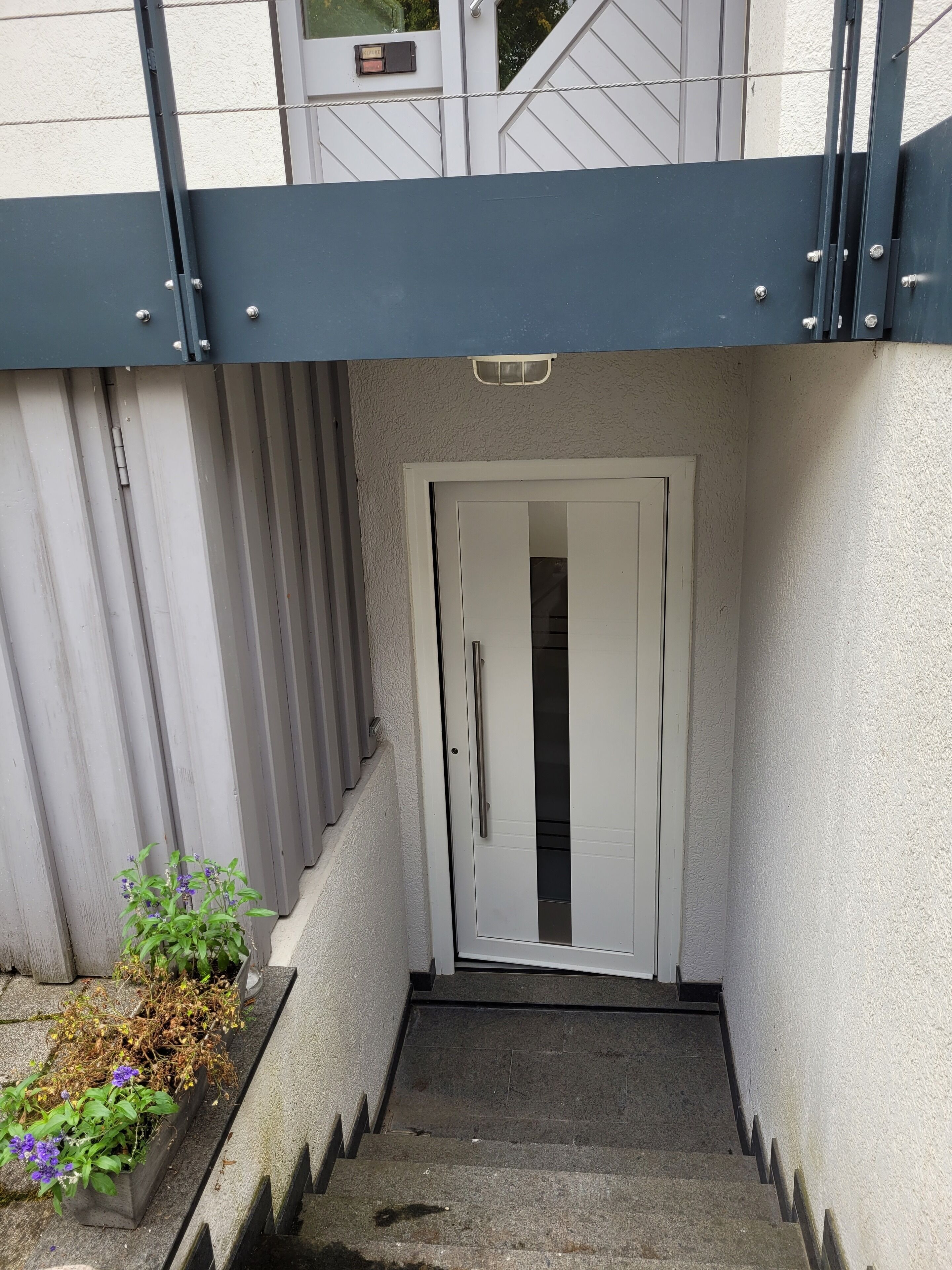 Una escalera estrecha que conduce a una puerta blanca con manija, flanqueada por barandas metálicas y una pequeña maceta con flores.
