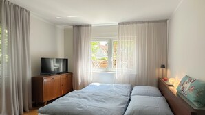 Une chambre à coucher avec un lit, un téléviseur posé sur un meuble en bois et une fenêtre avec des rideaux.