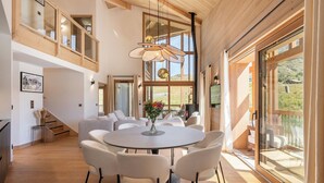 A spacious dining area with a round table and chairs, large windows offering a view of the outdoors, and a wooden ceiling.