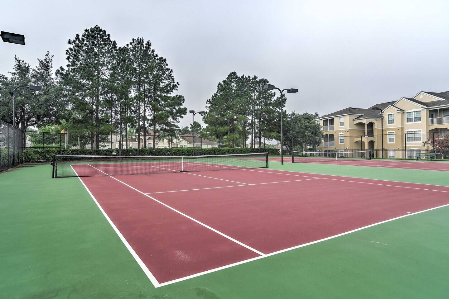 Un court de tennis en plein air, doté d’un filet, entouré d’arbres et avec un bâtiment visible à l’arrière-plan.