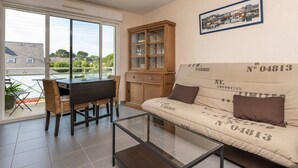 Ein Wohnzimmer mit Sofa, Glastisch, Esstisch mit Stühlen, Schrank und einem großen Fenster.