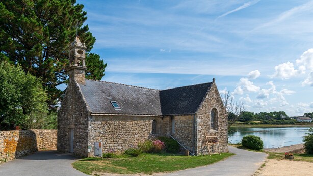 Eine Steinkirche mit Glockenturm, einem gepflasterten Weg und einem See im Hintergrund.