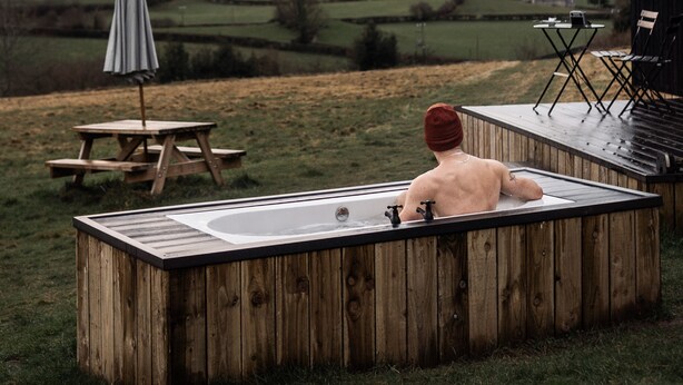 A wooden outdoor hot tub with a table and chairs, set against a rural landscape with fields and hills.