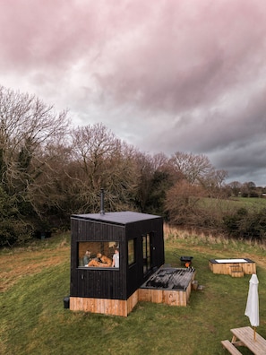 A modern black cabin with a large window, surrounded by a grassy area with wooden benches and a white umbrella. - Family Friendly Off-Grid Cabin In North Wales (Holywell)