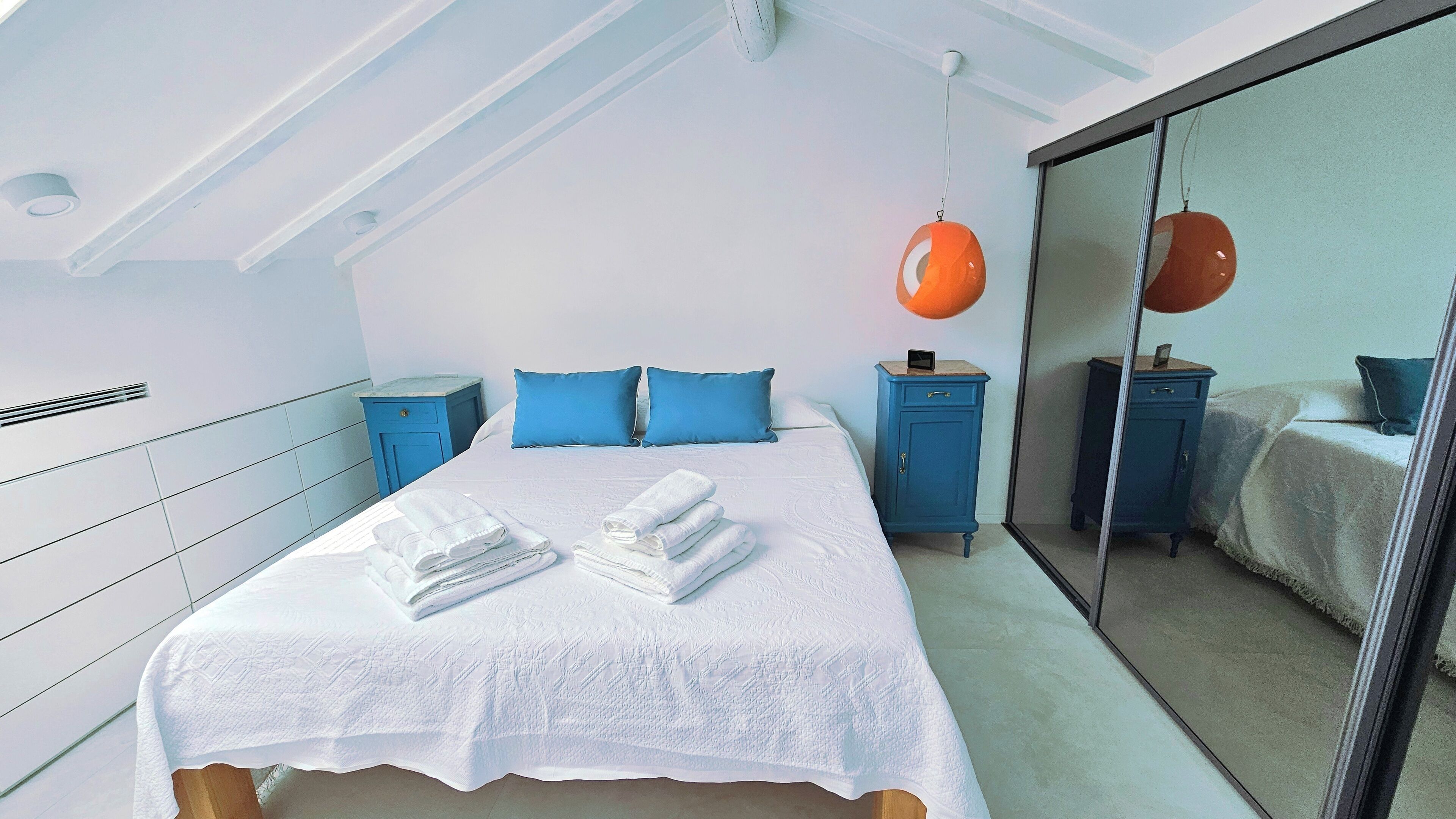 Een slaapkamer met een groot bed, twee blauwe nachtkastjes, een spiegel en een plafondventilator.