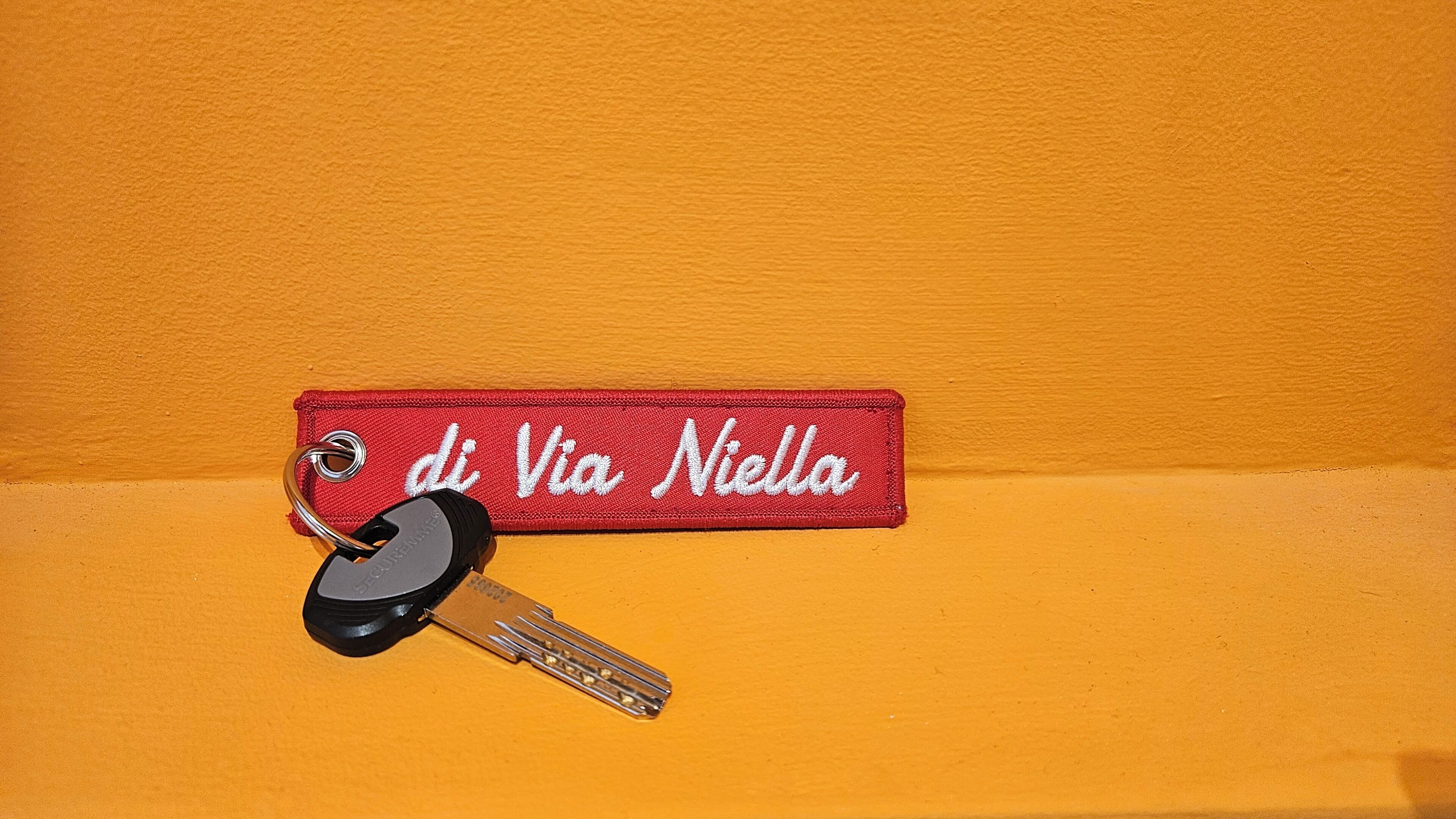 Ein Schlüssel mit einem roten Schlüsselanhänger mit der Aufschrift 'di Via Nella' an einer orangefarbenen Wand.