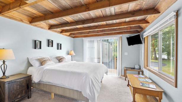 A bedroom with a wooden ceiling, a bed, a nightstand, a table, a TV, and a window with a view of trees. -  Scenic riverfront cottage with boat landing and docks. (Nekoosa)