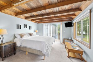 A bedroom with a wooden ceiling, a bed, a nightstand, a table, a TV, and a window with a view of trees.