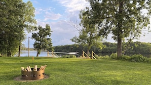 A grassy park area with a fire pit, wooden deck, and a lake in the background. -  Scenic riverfront cottage with boat landing and docks. (Nekoosa)