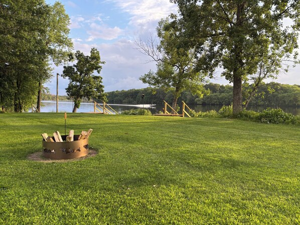 A grassy park area with a fire pit, wooden deck, and a lake in the background.