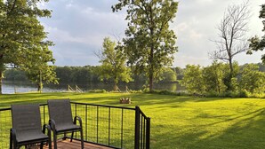 A deck with two chairs and a table, overlooking a grassy area with trees and a lake. -  Scenic riverfront cottage with boat landing and docks. (Nekoosa)