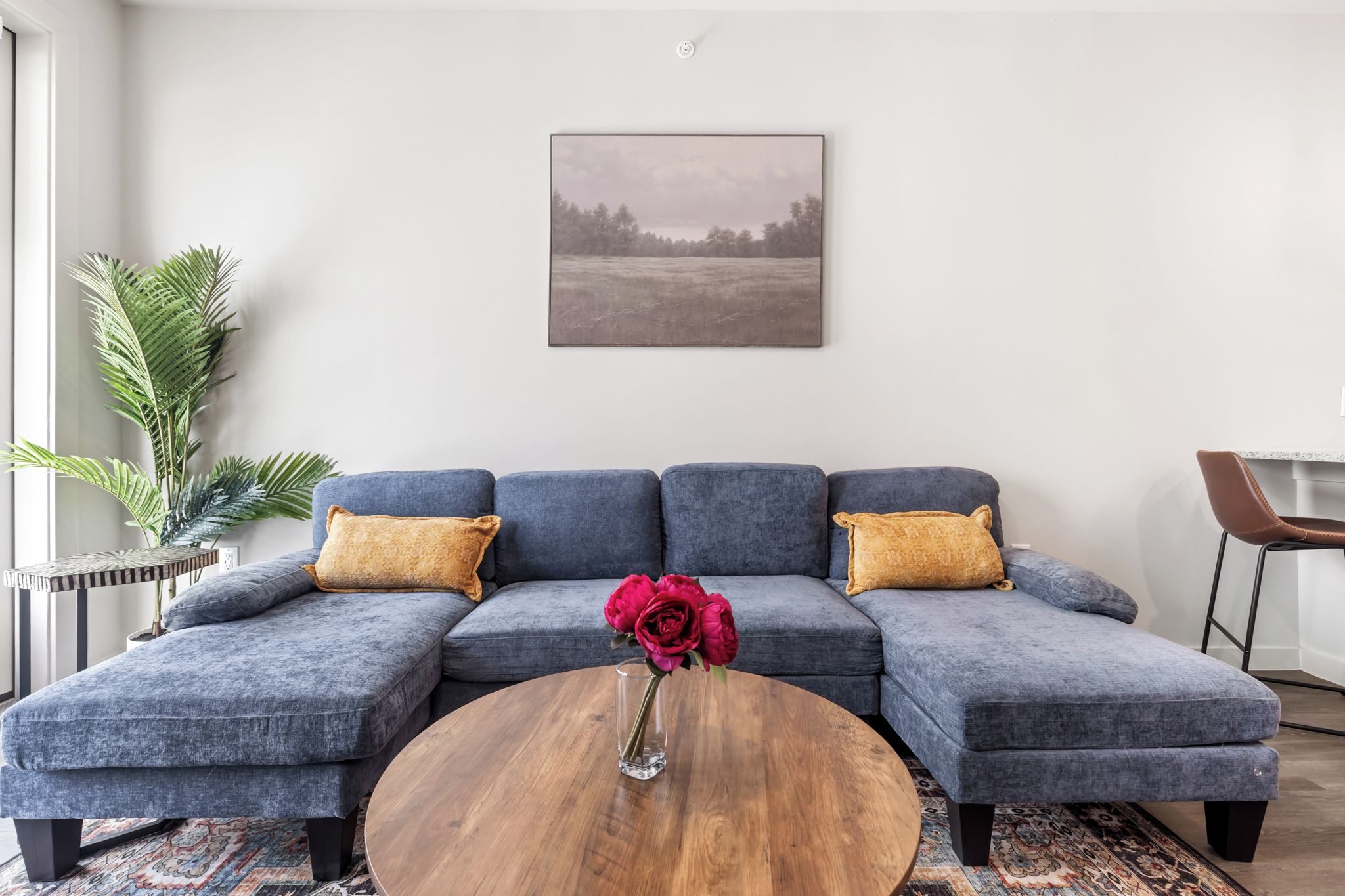 Un salon moderne avec un canapé d’angle, une table basse ronde en bois et un tableau représentant un paysage, accroché au mur.