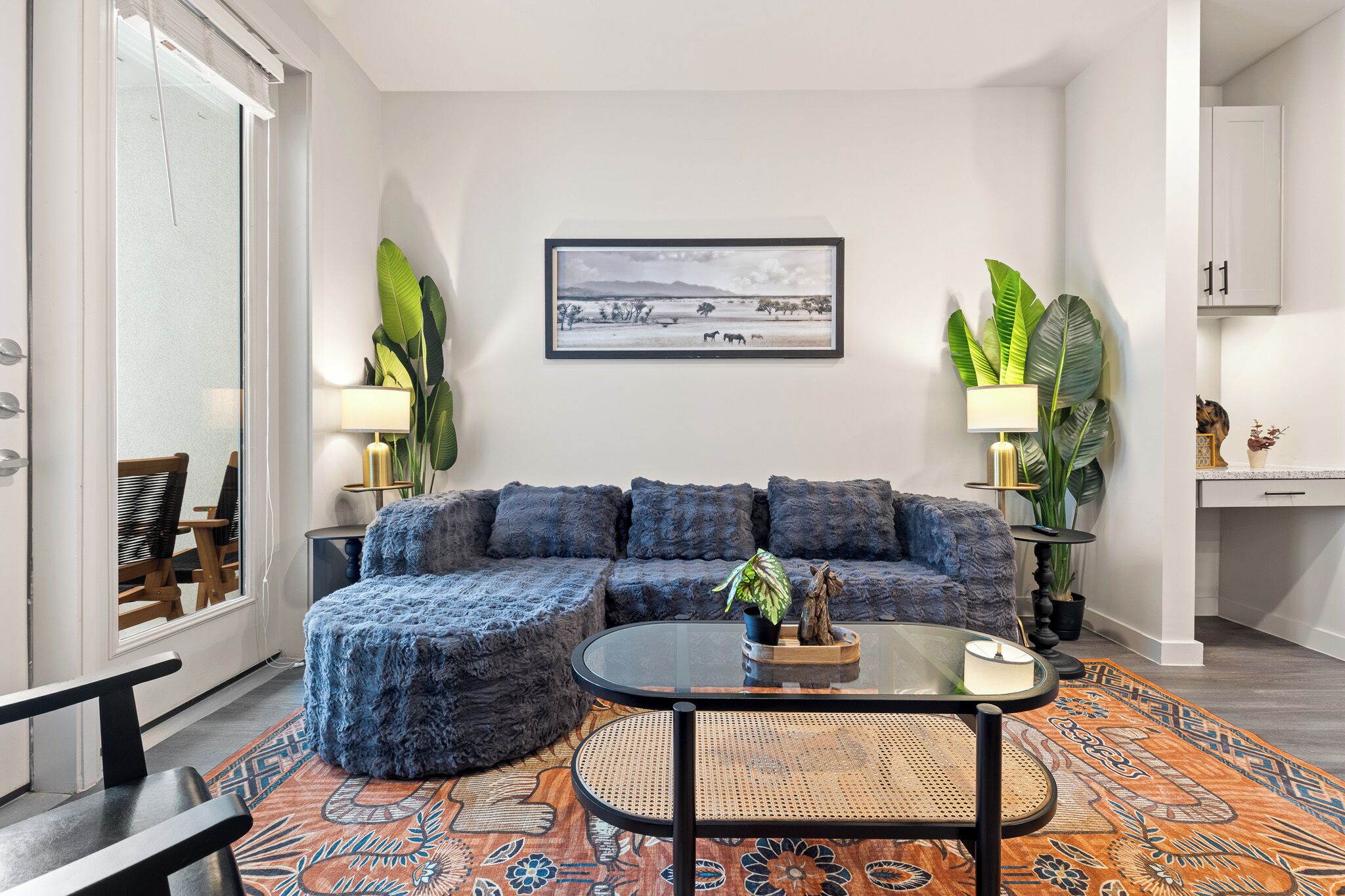 Ein Wohnzimmer mit einer Couch, Hocker, Couchtisch und zwei Lampen. An der Wand hängt ein gerahmtes Bild und links befindet sich eine Tür.