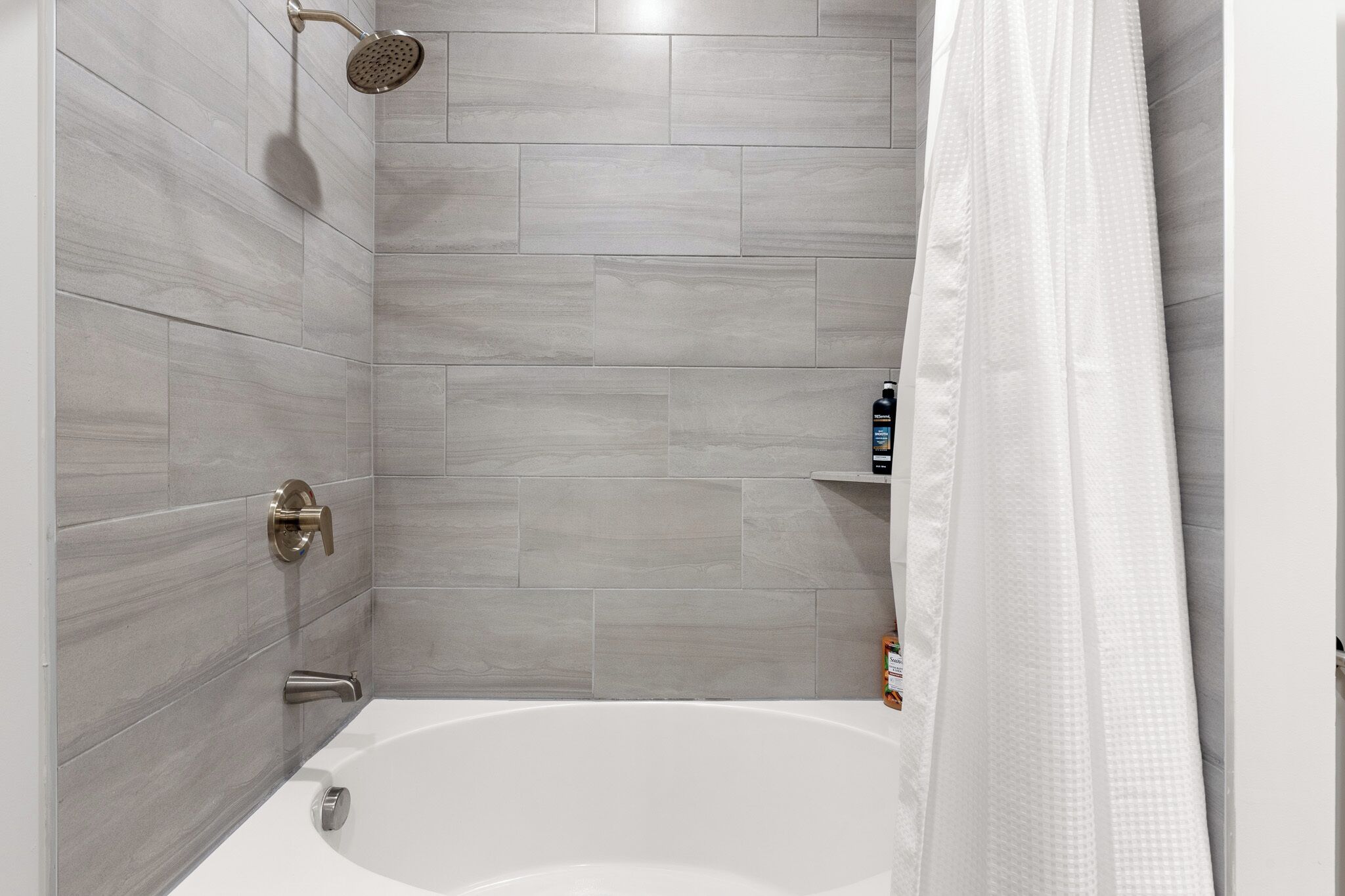 A modern bathroom with grey tiles, a white bathtub, a shower head, and a white shower curtain.