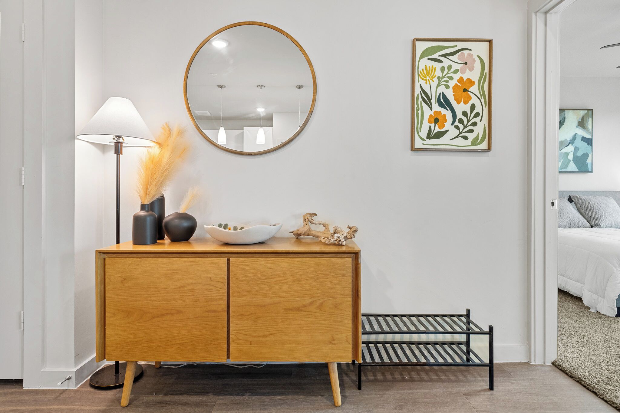 Une chambre moderne avec une commode en bois, un miroir rond et un lit recouvert d’une couette blanche.