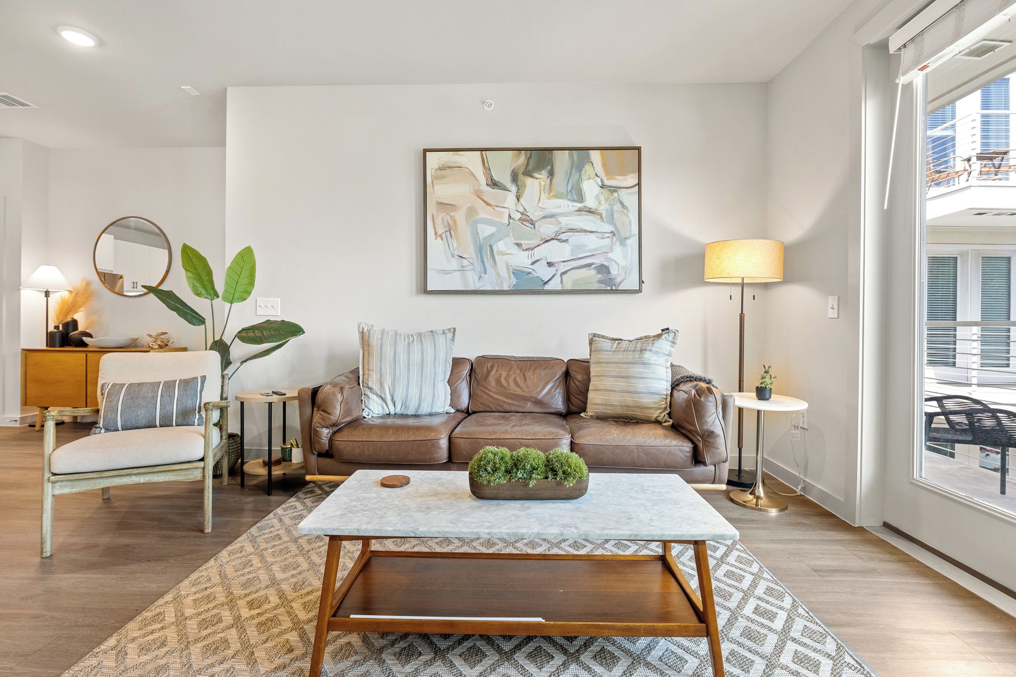 A modern living room with a brown leather sofa, a coffee table with a plant, a large abstract painting, and a view of the city through a large window.