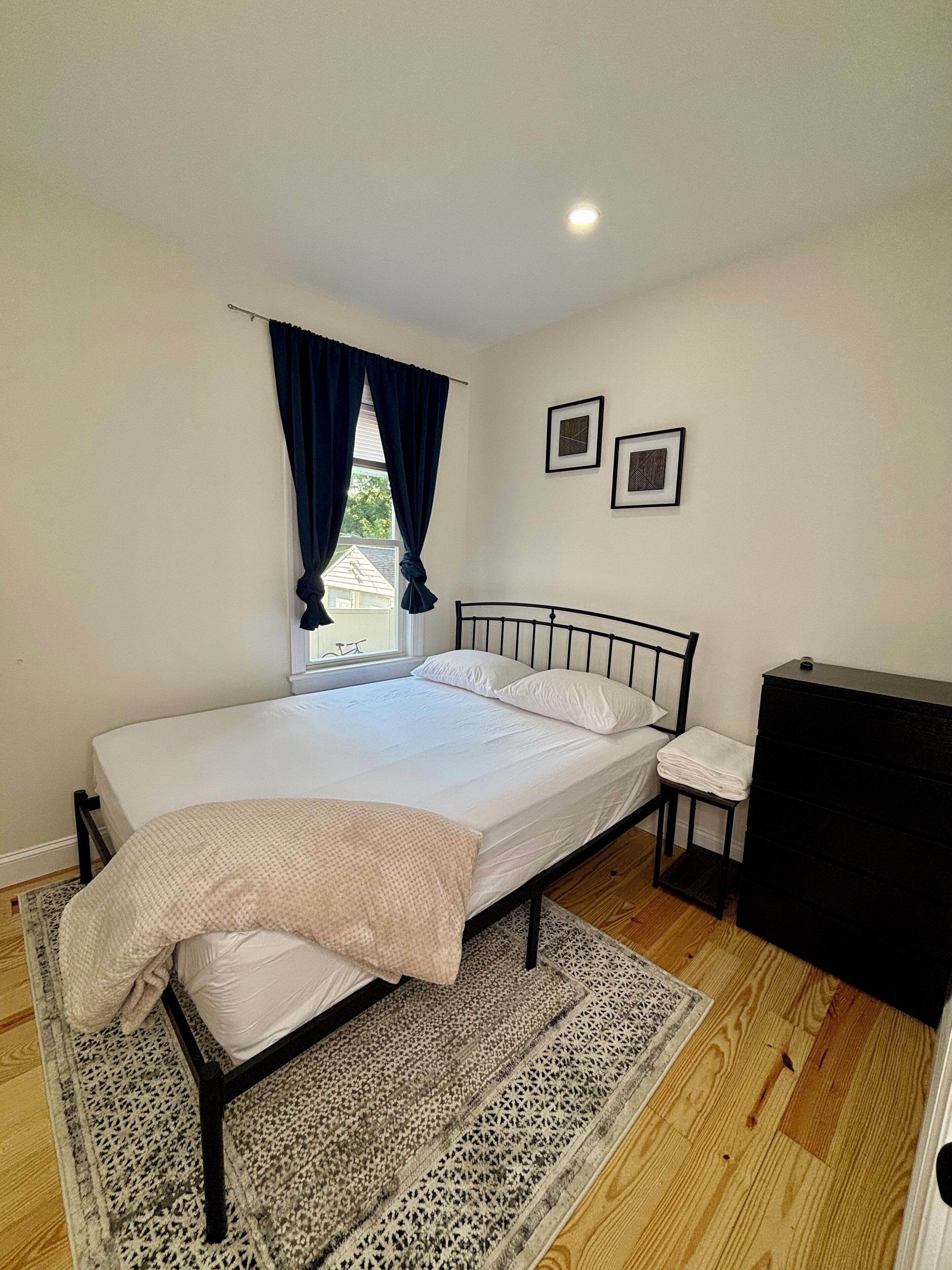 Une chambre bien rangée, avec un lit, une table de chevet, une commode et une fenêtre avec des rideaux.