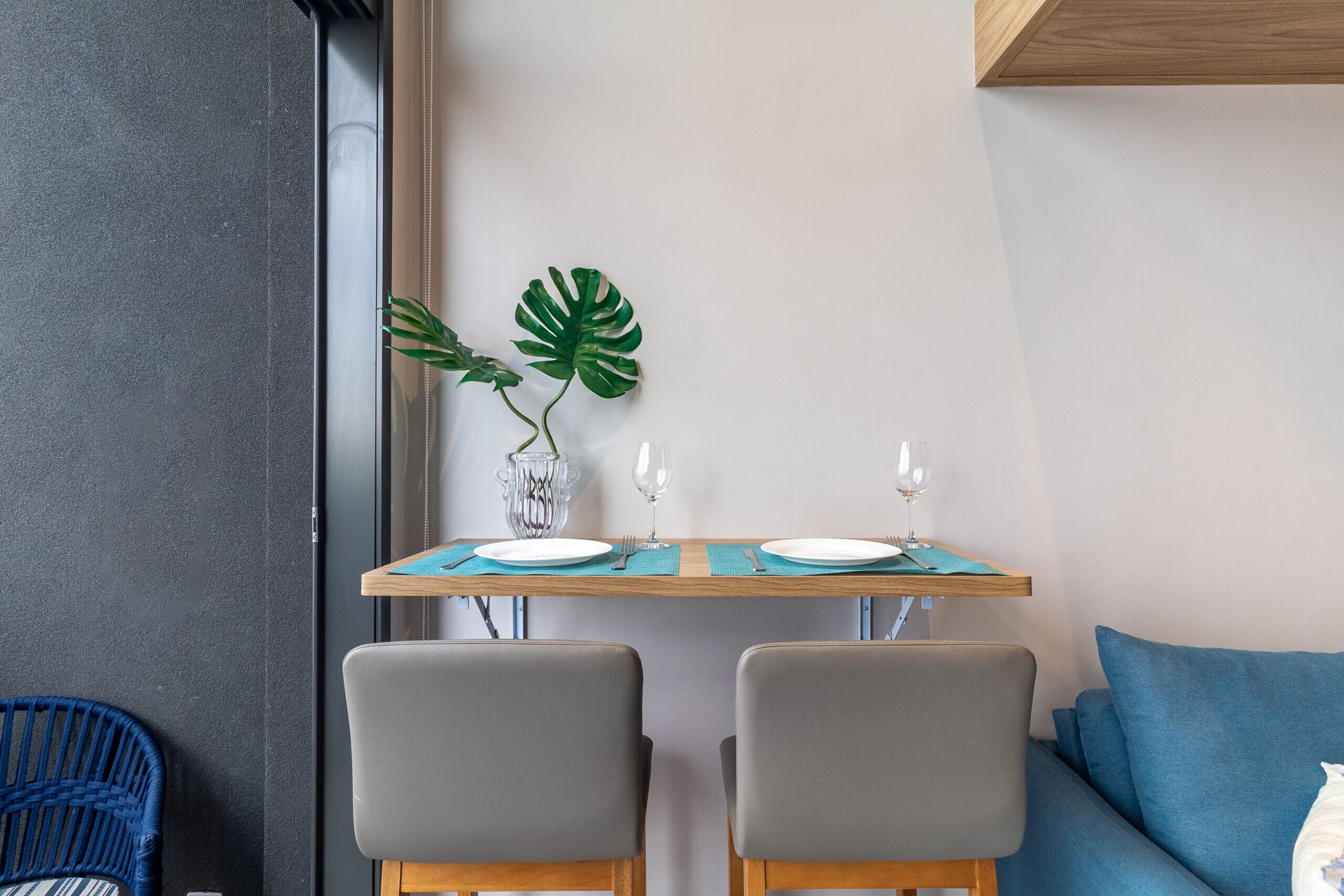 Un área de comedor con una mesa dispuesta para dos personas, dos sillas grises, un sofá azul y una planta verde grande sobre la mesa.