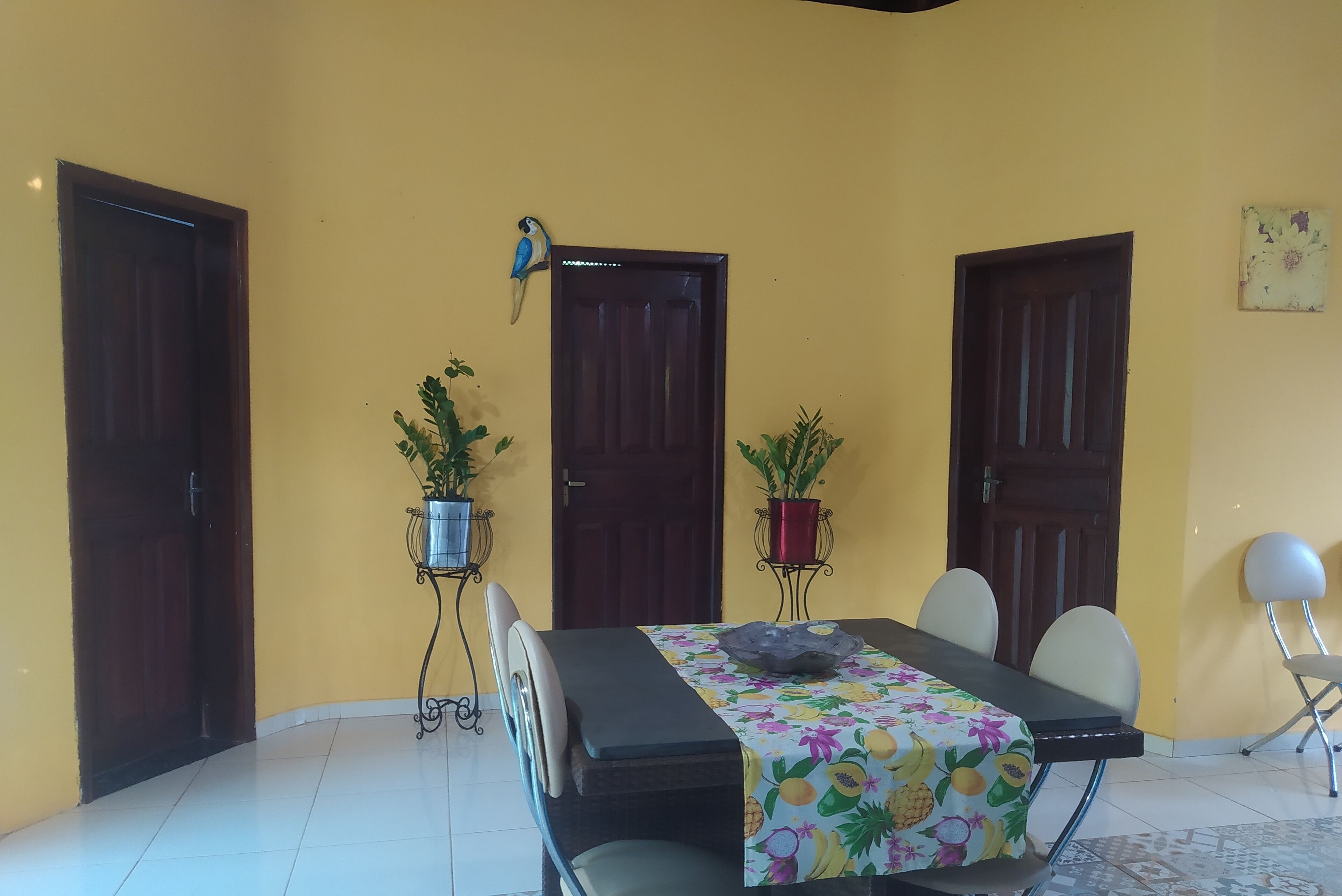 A dining area with a table covered in a fruit-patterned tablecloth, surrounded by chairs. There are two potted plants on metal stands, and a wall-mounted bird sculpture.