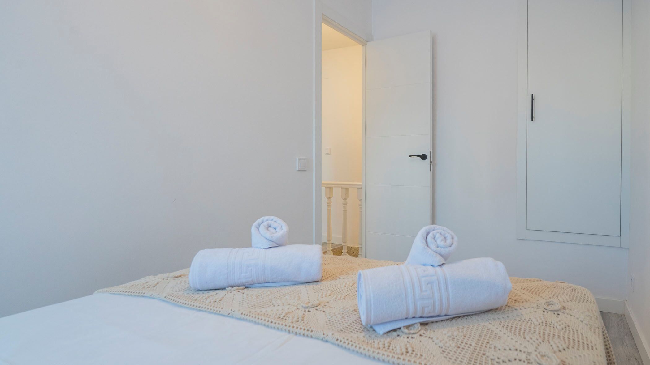 A bedroom with a bed, two rolled towels, and a door leading to another room.