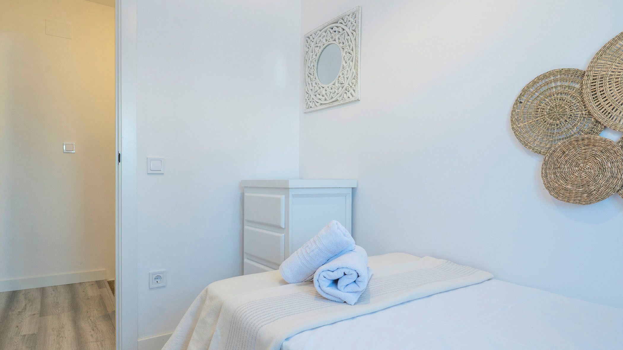 Ein schlicht eingerichtetes Schlafzimmer mit einem ordentlich bezogenem Bett, einem dekorativen Spiegel und Wandkunst aus gewebtem Material.