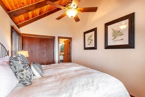 Une chambre avec un plafond en bois, un lit recouvert d’une courtepointe blanche, deux reproductions botaniques encadrées accrochées au mur et un ventilateur de plafond.