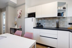 A modern kitchen with white cabinets, a black countertop, and a microwave. There is a dining table with chairs, a vase with dried flowers, and a picture of flowers on the wall.