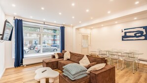Un salon moderne avec un canapé d’angle, un petit tabouret blanc, une table à manger en verre avec des chaises en métal et une grande fenêtre avec des rideaux.