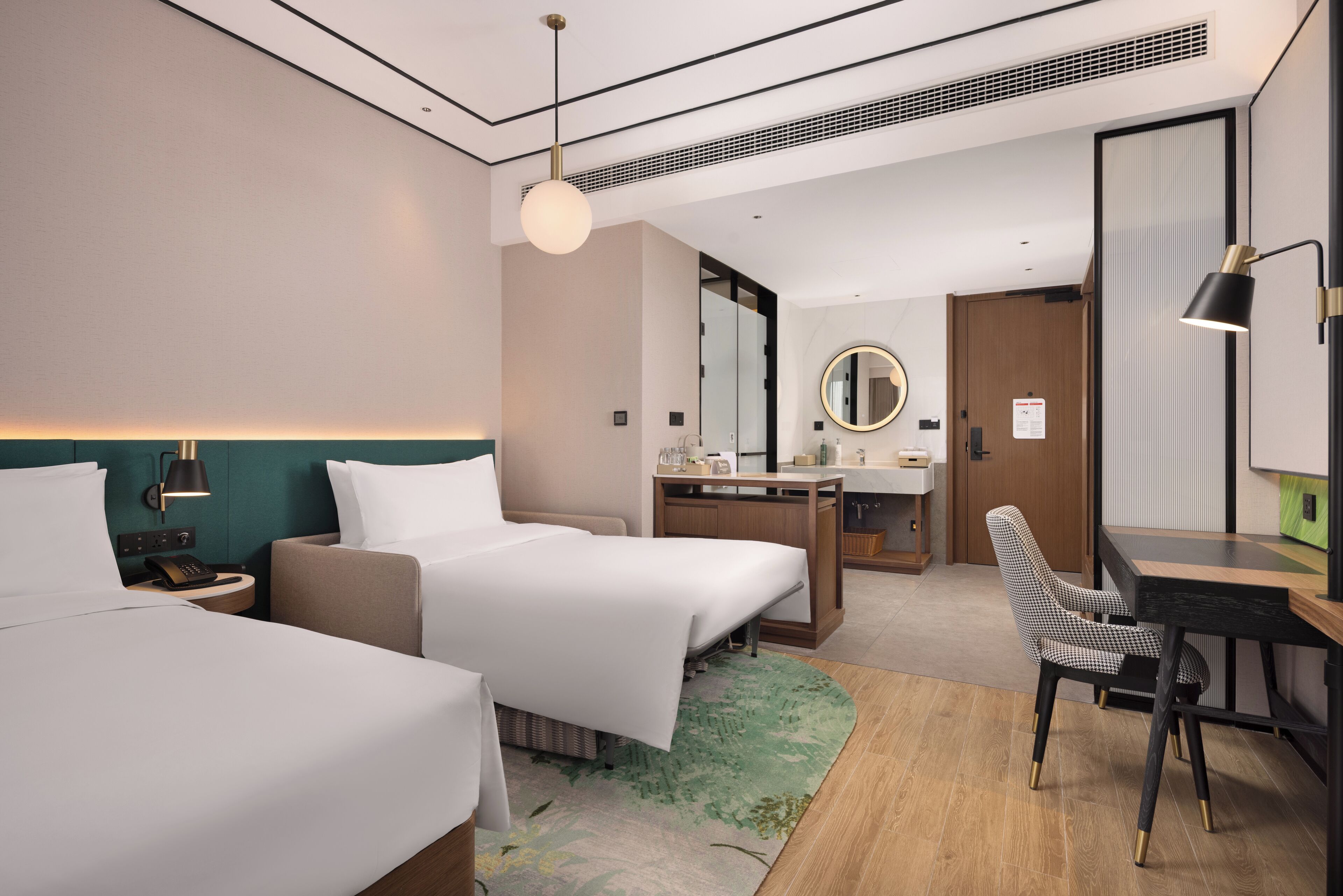A hotel room with two beds, a desk, a chair, a mirror, and a small kitchenette.