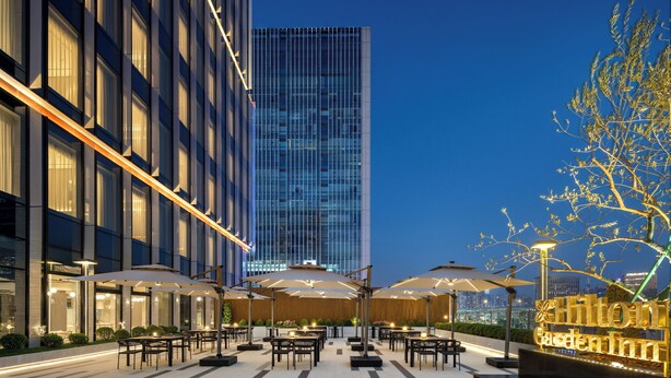 Un espace extérieur aménagé en terrasse, avec des tables et des parasols, un sol à carreaux et, en arrière-plan, un bâtiment dont les enseignes sont illuminées.
