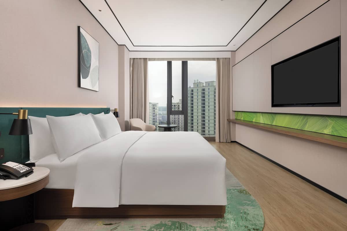 Ein Hotelzimmer mit einem großen Bett, einem Schreibtisch mit Telefon, einem an der Wand befestigten Fernseher und Blick auf hohe Gebäude durch das Fenster.