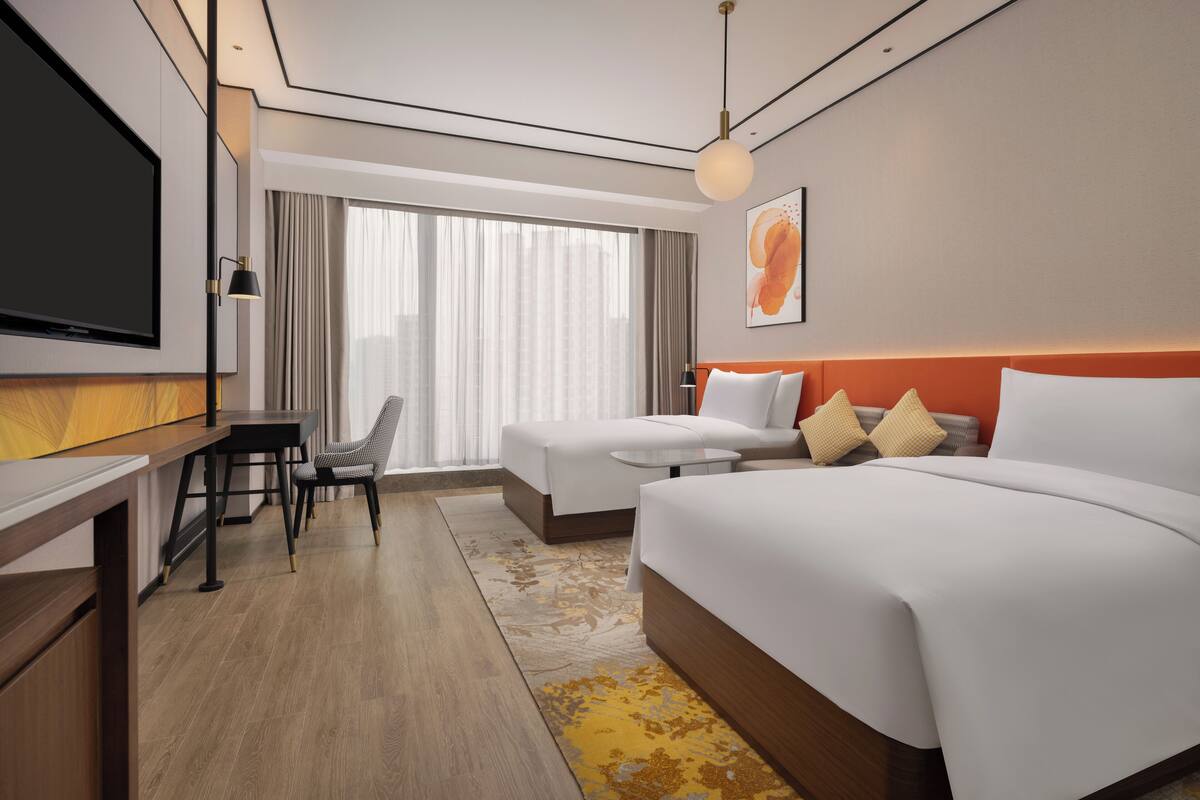 Ein Hotelzimmer mit einem großen Bett, einem Schreibtisch mit Stuhl, einem Fernseher und einem großen Fenster mit Vorhängen.