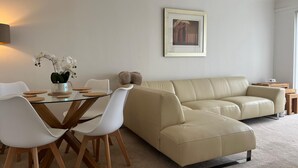 Una sala de estar moderna con una mesa de comedor de vidrio, sillas blancas, un sofá color beige y un cuadro en la pared.