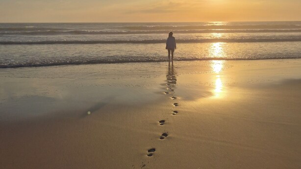 Una playa con huellas que conducen al océano, una persona de pie mirando la puesta de sol.