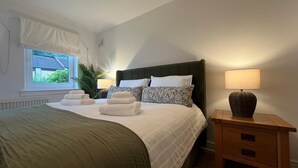 Una cama bien hecha con sábanas blancas y un plaid verde, una mesita de noche con una lámpara, y una ventana con vista a follaje.