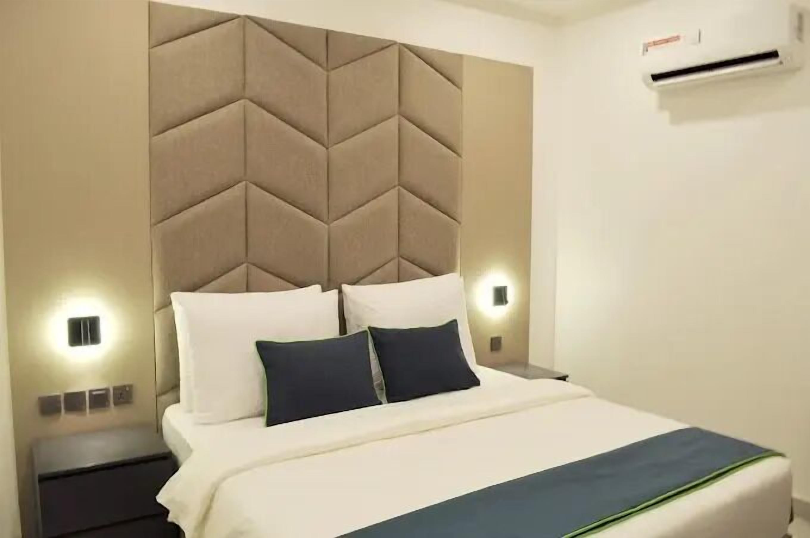A hotel room with a large bed, a padded headboard, bedside lamps, and an air conditioning unit.