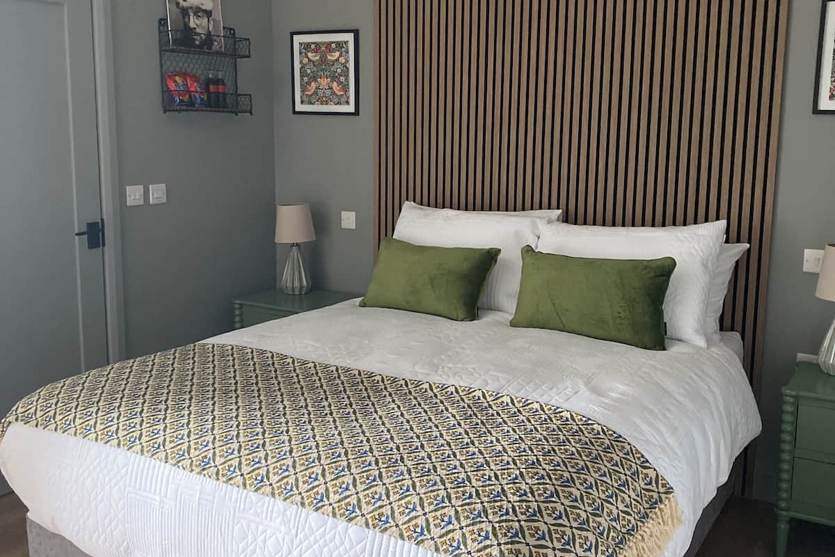 A bedroom with a bed, green pillows, a patterned bedspread, a wall-mounted shelf with items, and a striped headboard.